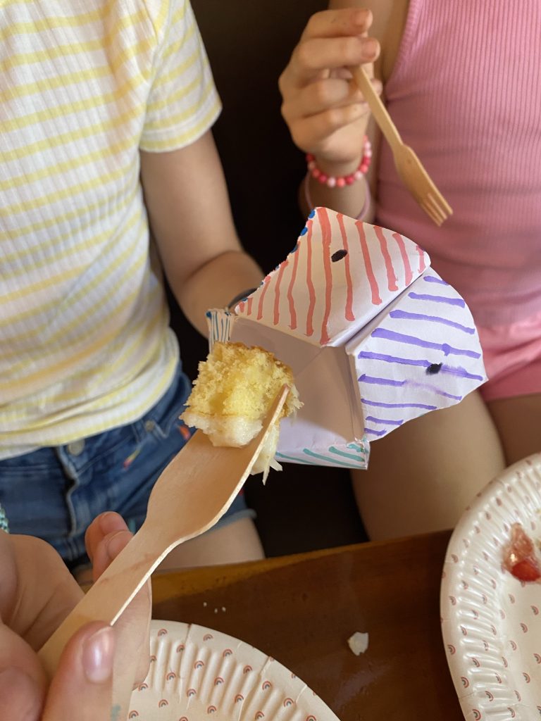 Kind isst ein Stück Kuchen mit einer Holzgabel, daneben ein selbst gebasteltes Himmel-und-Hölle-Papiertierchen