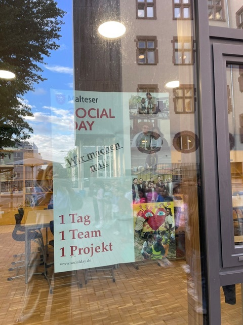 Plakat des Malteser Social Day mit dem Motto „Wir machen das! 1 Tag, 1 Team, 1 Projekt“ hinter einer Glastür der Mühlbergschule