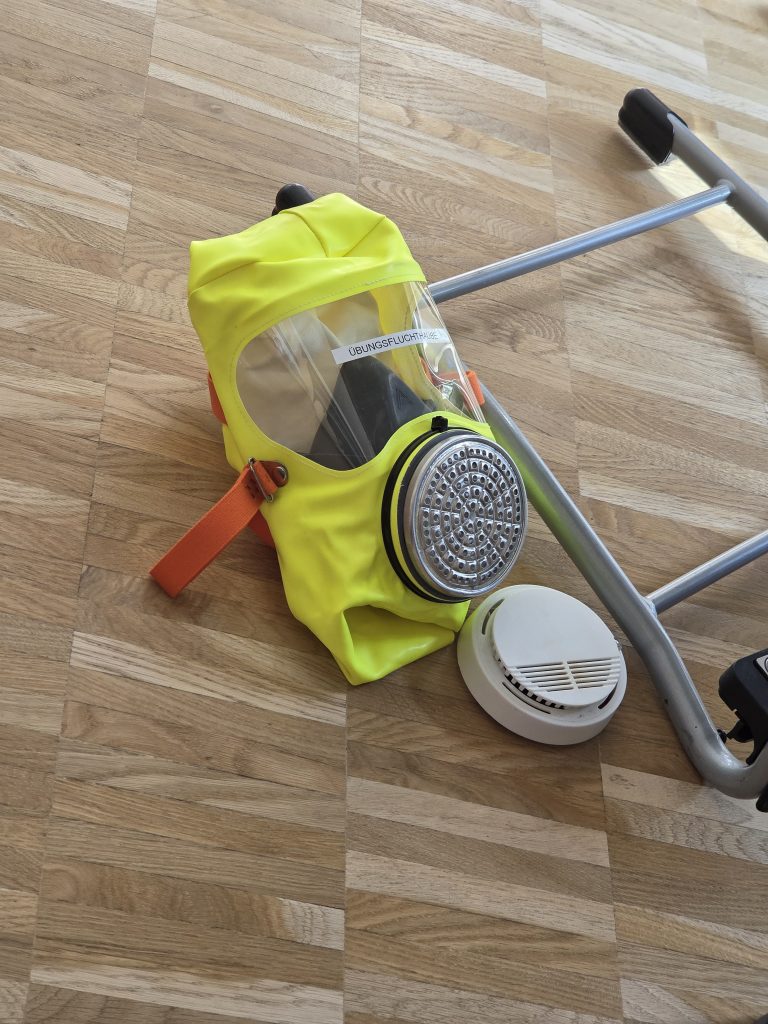 Gelbe Fluchthaube mit Atemfilter neben einem Rauchmelder auf dem Boden, Brandschutzerziehung Jahrgang 4