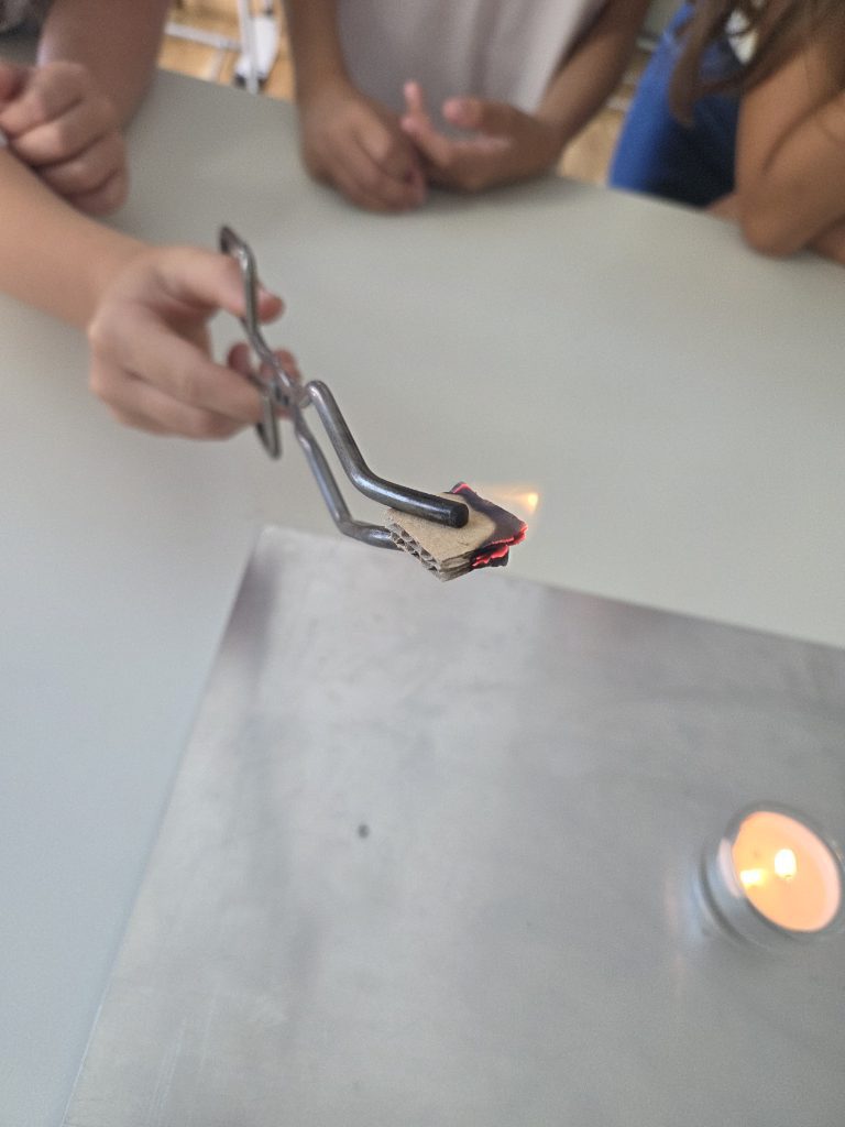 Hand hält mit einer Zange ein brennendes Papierstück über einer Kerze, Feuerversuch der Brandschutzerziehung
