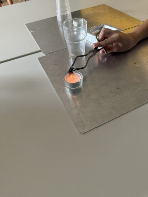Zange hält Material in die Teelichtflamme auf einer Metallplatte, Feuerversuch in der Brandschutzerziehung
