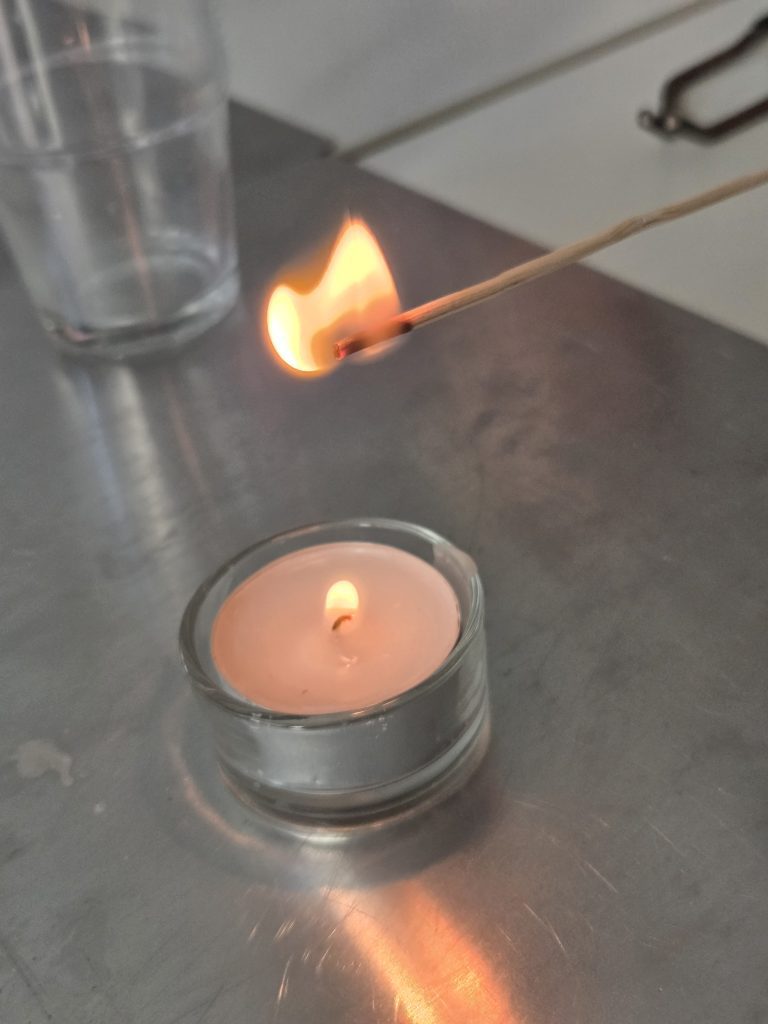 Brennendes Streichholz neben einem Teelicht auf einer Metallplatte, Feuerversuch der Brandschutzerziehung