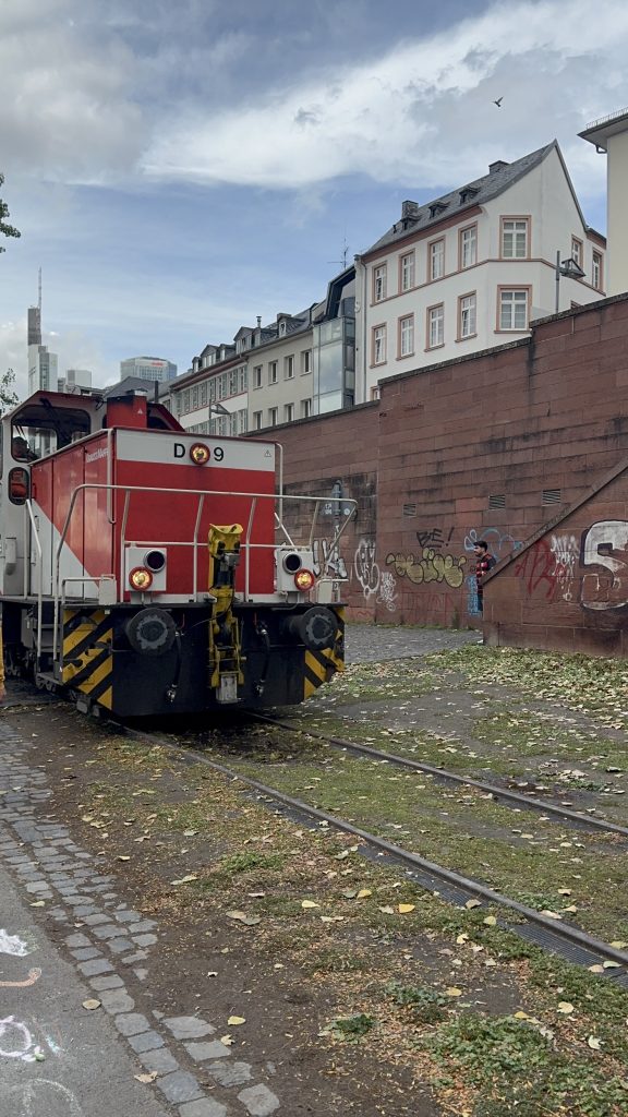 Rote Rangierlok auf Gleisen vor einer Backsteinmauer und Häusern am Mainufer in Frankfurt