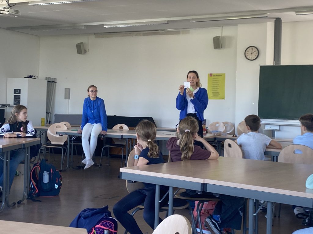 Zwei Frauen in blauen Jacken erklären im Klassenraum, Kinder hören zu, Mathematikworkshop Schillerschule