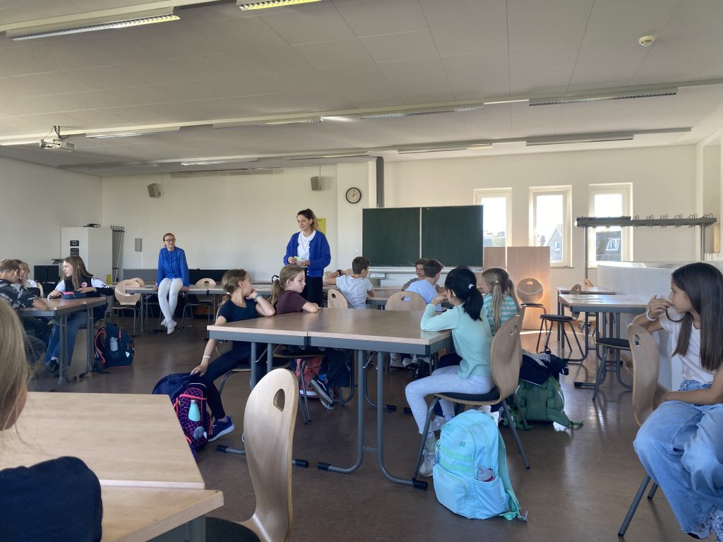 Kinder arbeiten in Gruppen an Tischen, zwei Leiterinnen im Hintergrund, Mathematikworkshop Schillerschule